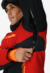 Giacca da sci rossa e nera con dettagli arancioni. Presenta una cerniera sotto il braccio e un rivestimento interno testurizzato nero. Foro per il pollice nei polsini.