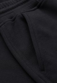 Gros plan sur un tissu noir texturé avec une poche latérale et une ceinture élastique, probablement partie d'un pantalon ou d'un short décontracté.