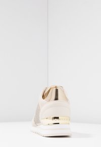 Beige sneaker with textured suede and perforated mesh. Features a metallic gold accent at the heel and a white sole with subtle stripes.