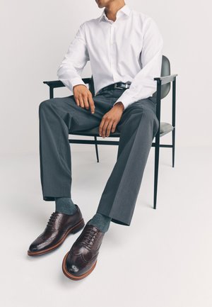 Homme portant une chemise blanche, un pantalon gris, des chaussettes bleues et des chaussures brogue marron assis sur une chaise minimaliste en métal noir.