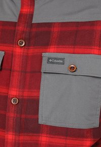 Chemise à carreaux rouges et rouge foncé avec poches poitrine grises boutonnées, boutons marron et étiquette logo Columbia sur le rabat de la poche droite.