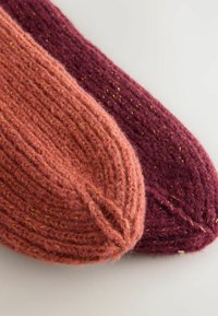 Zwei gestrickte Wollsocken, eine in staubigem Rosa mit goldenen Sprenkeln, die andere in tiefem Burgunderrot, mit geripptem Muster und abgerundeten Zehen.