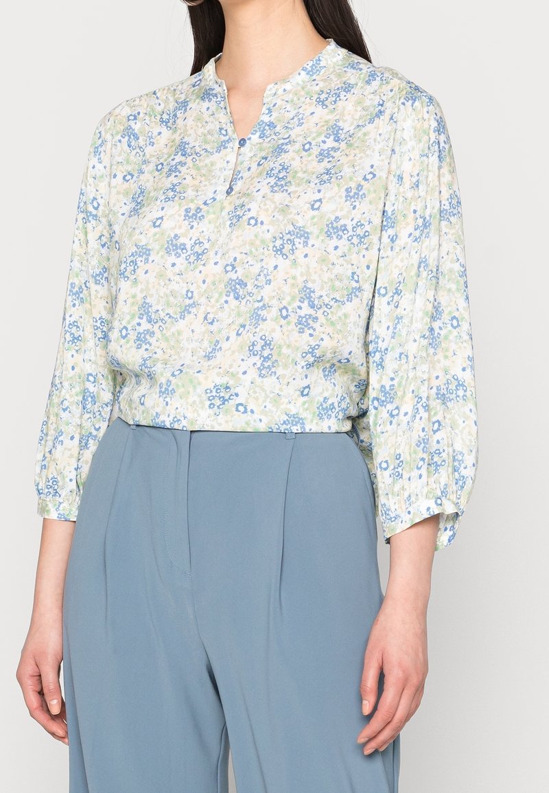 Femme portant un chemisier léger à fleurs bleues et vertes avec des manches trois-quarts bouffantes, rentré dans un pantalon bleu taille haute.