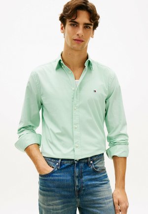 Młody mężczyzna z podwiniętymi rękawami, ubrany w jasnozieloną koszulę zapinaną na guziki i niebieskie dżinsy, stojący z jedną ręką w kieszeni.