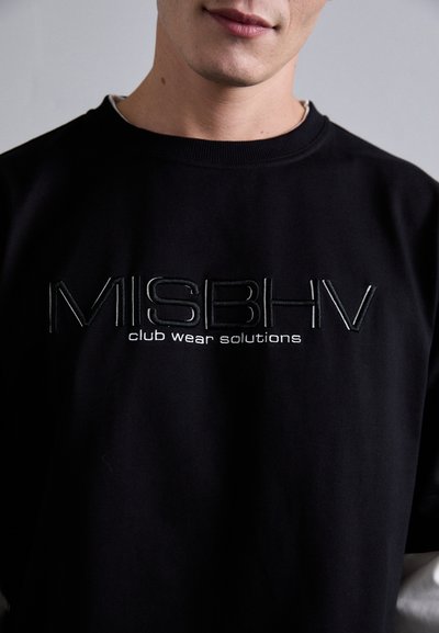 Mees, kes kannab musta kapuutsiga särki, millel on rinnal ingliskeelne kiri "MISBHV club wear solutions", taustal lihtne taust.