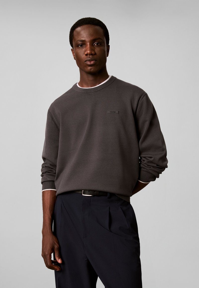 Jeune homme portant une chemise à manches longues gris foncé et un pantalon noir, debout devant un fond uni gris clair.
