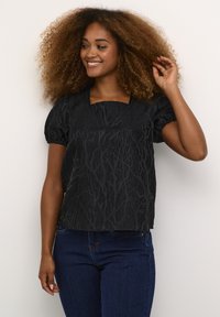 Zwarte top met een getextureerd takkenpatroon, vierkante halslijn en pofmouwen, gecombineerd met donkerblauwe jeans. Gladde stof met een subtiele glans.