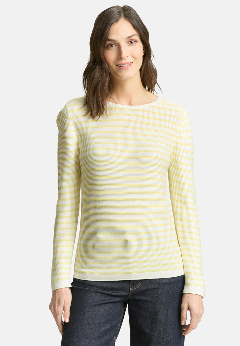 Femme aux cheveux châtains mi-longs portant un pull rayé blanc et jaune à manches longues et un jean bleu foncé, debout devant un fond blanc.