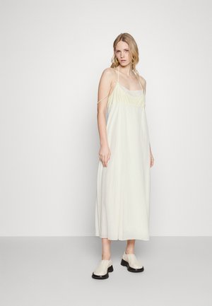 Robe maxi sans manches de couleur jaune clair en tissu doux avec des bretelles fines et une coupe décontractée, assortie à des chaussures à talons épais de couleur claire.