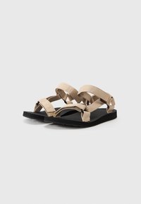 Beige sandaler med kryssede elastiske stropper, flat svart gummisåle og teksturert såle for grep. Minimalistisk design, åpen tå stil.