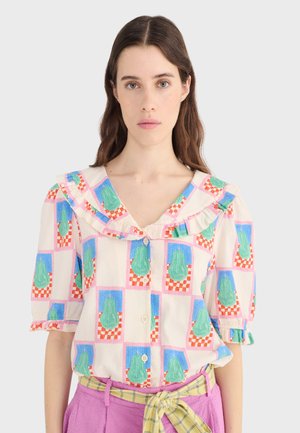 Kobieta o długich brązowych włosach ubrana jest w kolorową koszulę w kratę z zielonymi nadrukami gruszek oraz różowe spodnie przewiązane żółtym paskiem w kratę.