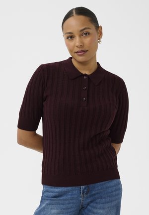 Nő sötétlila rövid ujjú kötött galléros pólóingben, gombokkal, kék farmerral, egyszínű fehér háttér előtt pózolva.