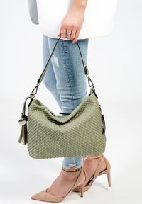 Zielona pleciona torba na ramię o luźnym kroju, z detalem w postaci frędzla i srebrnymi akcentami, w połączeniu z jasnoniebieskimi dżinsami i cielistymi szpilkami.