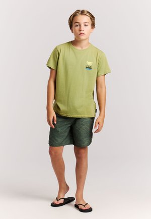 Green cotton t-shirt with short sleeves and small logo. Paired with patterned dark green shorts and black flip-flops.