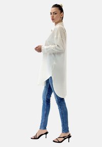 Camicia bianca a maniche lunghe con orlo leggermente curvo, abbinata a jeans blu aderenti e tacchi neri con cinturini. Tessuto morbido, vestibilità moderna.
