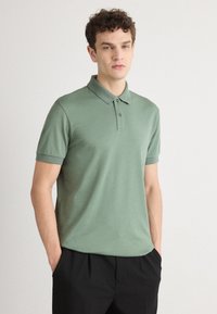 Grønt bomullspolo med krage, to-knappsklaff og korte ermer, vist på en modell med hender i lommene, med diskret logodetalj.