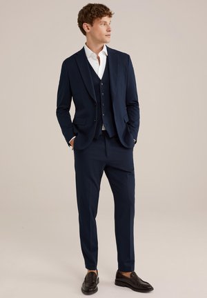 Marinblå tre-delad kostym med en skräddarsydd design, som inkluderar en kavaj, väst och skräddarsydda byxor, tillsammans med en vit skjorta och svarta loafers.