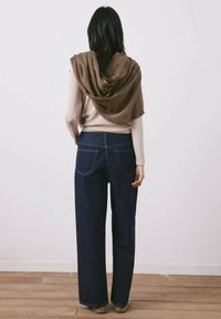 Persona con cabello largo y oscuro, que lleva un suéter claro, una bufanda marrón sobre los hombros y unos jeans oscuros de pierna ancha, de pie contra una pared lisa.