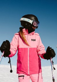 Růžová lyžařská bunda se zipem vpředu, černé rukavice a černá helma s světle modrým akcentem, proti jasné modré obloze.