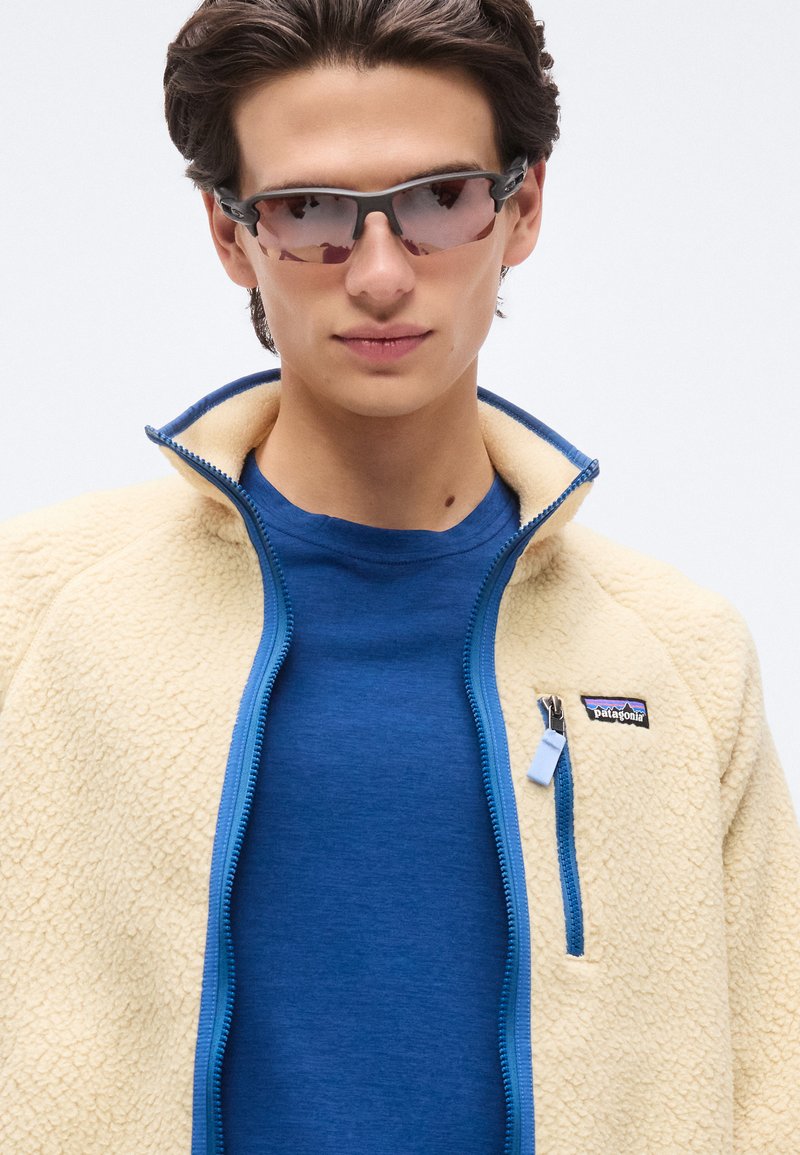 Jeune homme portant des lunettes de soleil sportives à verres teintés marron, une veste en polaire crème avec des bordures bleues, et un t-shirt bleu à col rond sur fond blanc.