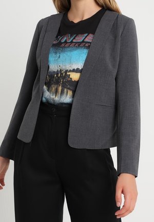 Kobieta ubrana w dopasowaną szarą marynarkę na czarnym T-shircie z grafiką i czarne spodnie z wysokim stanem na neutralnym tle.