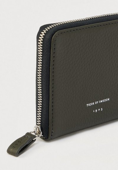 Σκούρο πράσινο δερμάτινο πορτοφόλι με υφή, ασημένιο φερμουάρ και λογότυπο με ανάγλυφη λευκή εκτύπωση. Διαθέτει στρογγυλεμένη λεπτομέρεια στο κράτημα του φερμουάρ.