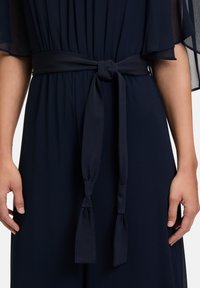 Marinfärgad klänning i tyg med en knytbar midjebälte med förlängda ändar, mjuka draperade ärmar och slät yta. Plisserad bystdetalj.