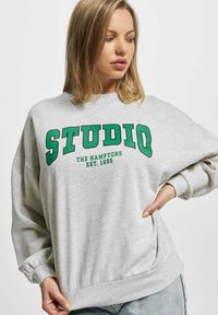 Grå oversized genser laget av bomull, med grønn tekst som sier "STUDIO THE HAMPTONS EST. 1995." Ribbet mansjetter og hem.
