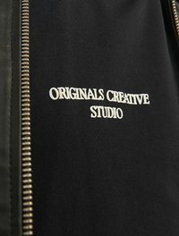 Jachetă neagră cu fermoar, dintr-un material texturat. Dispune de inscripția "STUDIO CREATIV ORIGINAL" brodată în alb și detaliu cu fermoar auriu pe față.