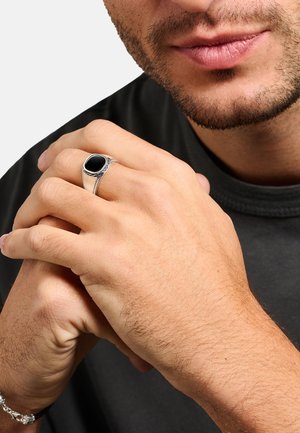 Mann mit kurzem Gesichtshaar, der ein schwarzes Hemd trägt, die Hände verschränkt haltend, zeigt einen silbernen Ring mit schwarzem Stein am Ringfinger der linken Hand.