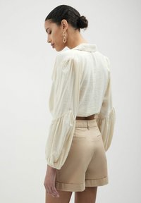 Blusa corta crema con maniche a sbuffo abbinata a pantaloni corti beige. La blusa presenta un colletto; i pantaloni hanno un orlo arrotolato e tasche posteriori.