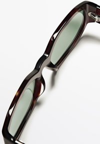 Sluneční brýle s tmavě vínovými, lesklými plastovými rámy a zelenými skly. Výrazný design se vyznačuje vlnitým tvarem a ostrými kovovými akcenty.