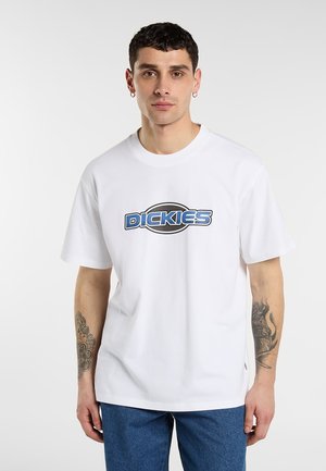 Man med kort mörkt hår som bär en vit Dickies t-shirt och blå byxor, visar tatueringar på båda underarmarna, stående mot en enfärgad bakgrund.