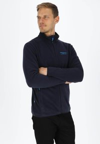 Marineblaue Fleecejacke mit hohem Kragen, Reißverschluss vorne und zwei Seitentaschen. Verfügt über blaue Akzente und ein gesticktes Logo auf der Brust.