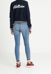 Námořnická bombarďácká bunda s bílým potiskem, modré úzké džíny a maroon tenisky Adidas se zlatými detaily. Neformální, přiléhavý silueta.