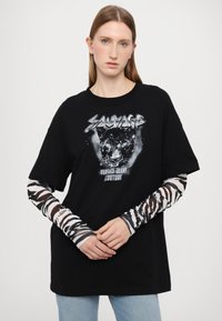 Svart oversized t-skjorte med hvit grafisk trykk som viser en panter og teksten "Salvage". Lange ermer med gjennomsiktige zebra mønsterdetaljer.