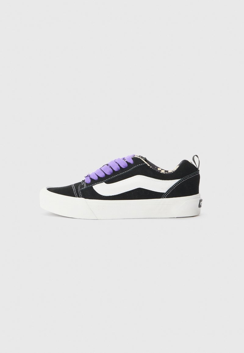 Baskets basses noires et blanches avec des lacets violets, une bande latérale blanche, une semelle épaisse blanche et une languette au talon, vues de côté sur fond gris.