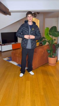 Juodas pūkinis švarkas su užtrauktuku, pilkas vidinis sluoksnis, derinamas su juodomis kelnėmis ir baltais sportbačiais. Vidaus aplinka su medinėmis grindimis ir augalais.