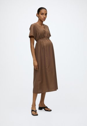 Femme enceinte en robe midi marron ample avec des boutons, debout pieds nus sur fond blanc, légèrement tournée vers la gauche.