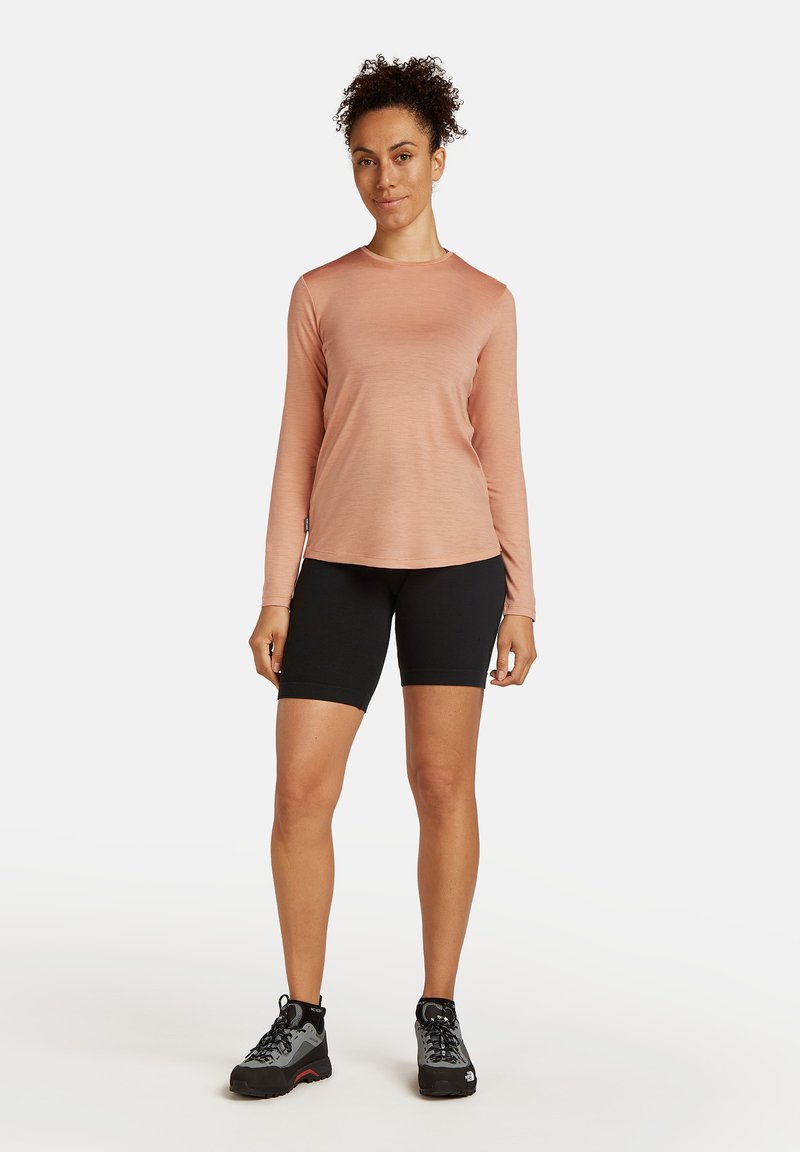 Langermige, rozenkleurige top in combinatie met zwarte fietsshorts. Model draagt grijze wandelschoenen met rode accenten. Glad, lichtgewicht materiaal.