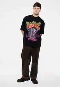 Black oversized t-shirt featuring multicolored graphic of a figure and text. Paired with dark brown corduroy pants and black shoes.