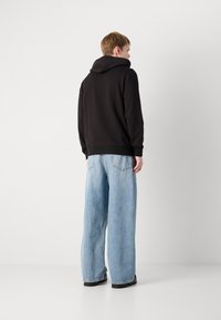 Persoon in een zwarte hoodie en lichtblauwe wijde jeans, staand tegen een effen witte achtergrond, met zijn rug naar de camera gekeerd.