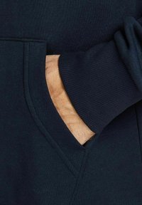 La felpa navy presenta una tasca a marsupio, polsini a costine e una morbida texture del tessuto. Ingrandimento della mano inserita nella tasca.