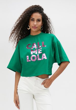 Camiseta corta verde de algodón, con gran texto en lentejuelas rosas "LLÁMAME LOLA", con mangas oversized y escote redondo.
