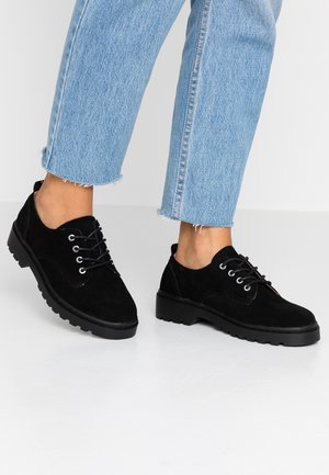 Czarne zamszowe buty na sznurówki z okrągłym noskiem, teksturowaną powierzchnią i gumową podeszwą. Noszone z postrzępionymi, jasnoniebieskimi dżinsami.