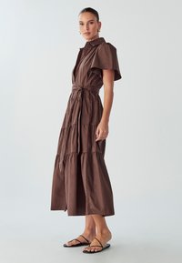 Rjava bombažna midi obleka s ovratnikom, kratkimi plaščnimi rokavi in večstopenjskim dizajnom. Ima pasni trak in stranski razporek.