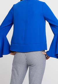 Jasnoniebieska, długosleeve bluzka z rozszerzonymi mankietami i zamkiem z tyłu. Połączona z czarno-białymi spodniami w kratkę. Gładka tekstura, dopasowany krój.