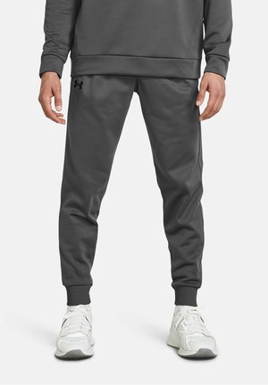 Mężczyzna ubrany w ciemnoszare spodnie dresowe Under Armour i białe buty sportowe, stojący na jednolitym tle.