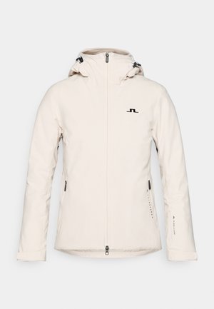 Veste à capuche beige avec fermeture éclair sur le devant, cordons de serrage ajustables sur la capuche, poches latérales zippées et logo noir sur la poitrine et la manche.