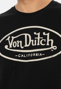 T-shirt en coton noir avec un grand logo blanc usé portant l'inscription "Von Dutch - CALIFORNIA" dans une police vintage audacieuse.